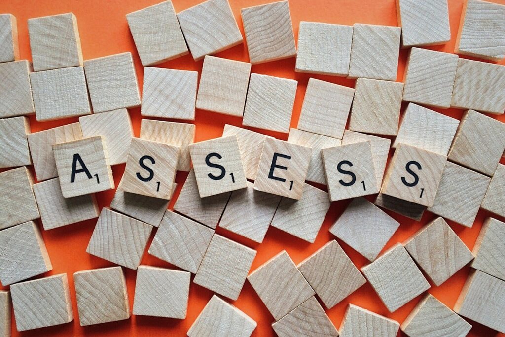 Wooden square blocks with letters on each one spell out the word "assess", while lying on a layer of blank blocks, all set against an orange backdrop.